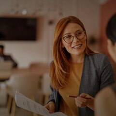 Eine Frau mit Brille und roten Haaren spricht lächelnd mit einer Kollegin und hält dabei ein Dokument in der Hand; im Hintergrund sitzt eine weitere Person an einem Tisch in einem modernen Büro.