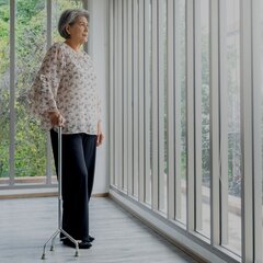 An elderly woman stands with a walking stick in a bright interior with floor-to-ceiling windows and moves independently through a barrier-free environment.