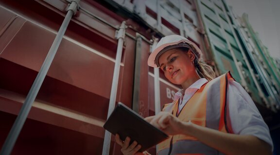 Young woman in a construction helmet and high-visibility waistcoat stands between stacked shipping containers and looks at a tablet.