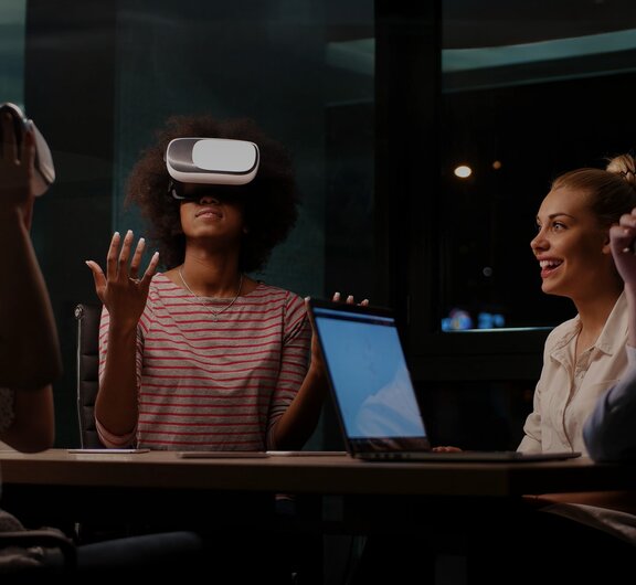 Eine Gruppe von Personen, die mit einem Laptop und Virtual-Reality-Brillen gemeinsam am Tisch sitzen und sich unterhalten.
