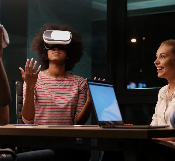 Eine Gruppe von Personen, die mit einem Laptop und Virtual-Reality-Brillen gemeinsam am Tisch sitzen und sich unterhalten. 
