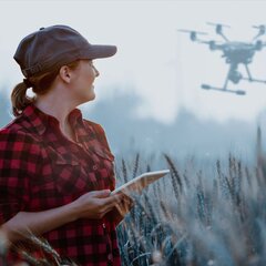 Eine Frau in kariertem Hemd und Kappe hält ein Tablet vor einer Drohne.